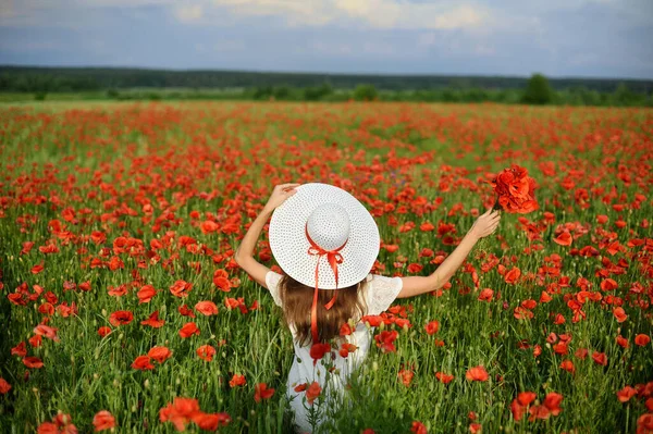 Красивая молодая женщина живой стиль держит в руках белую шляпу и стоит с распростертыми объятиями на поле красных маков на фоне синего неба и белых облаков. Вид сзади — стоковое фото