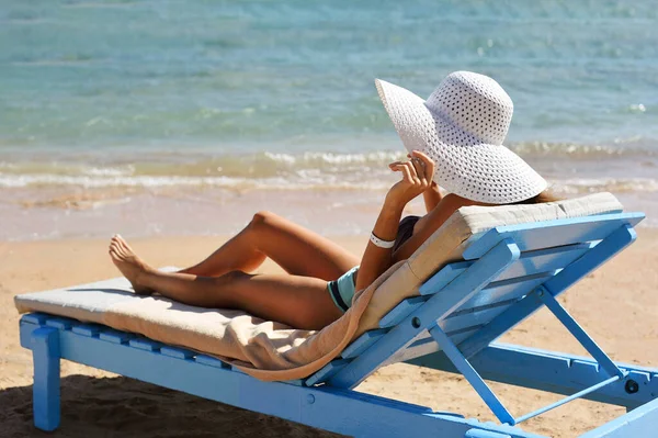 Piękna kobieta opalająca się na plaży w tropikalnym kurorcie turystycznym, ciesząca się wakacjami. Dziewczyna trzyma kapelusz z rękami na leżaku w pobliżu morza — Zdjęcie stockowe