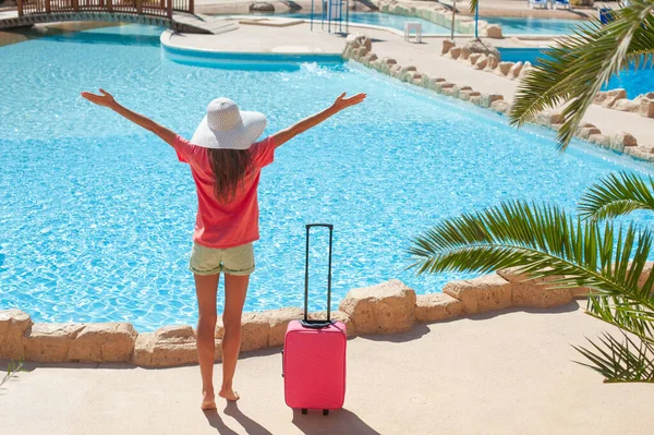 Viagens, férias de verão e conceito de férias - Mulher bonita levanta as mãos perto da área da piscina do hotel com mala rosa — Fotografia de Stock