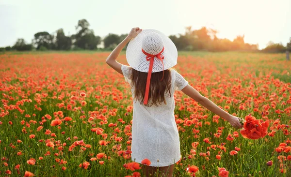Красивая молодая женщина живой стиль держит в руках белую шляпу и стоит с распростертыми объятиями на поле красных маков на фоне синего неба и белых облаков. Вид сзади — стоковое фото