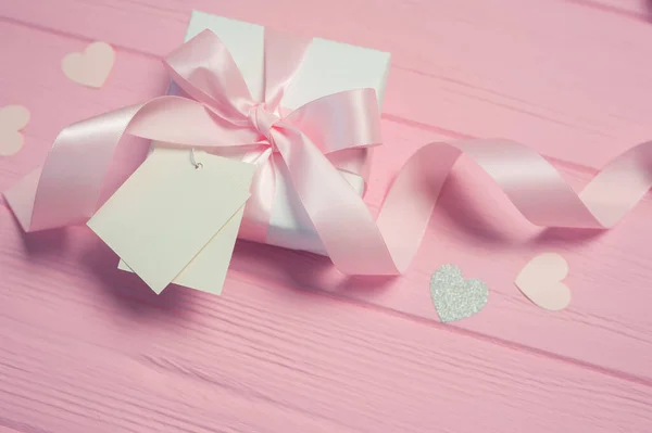 Caja de regalo blanca con lazo de satén rosa y cinta sobre fondo de madera rosa. La etiqueta de la maqueta de la boda se incluye para su texto. Puede utilizar para el día de San Valentín, día de la madre, cumpleaños — Foto de Stock