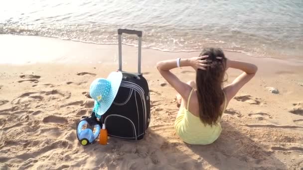 Beautiful woman sitting with a suitcase on the sunny beach and looking at the sea. Full HD video motion footage — Stock Video
