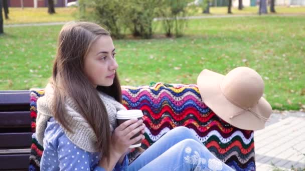 Mooi meisje zittend op een bankje in een zonnig park in de herfst. Het meisje drinkt warme koffie en buiten. jonge brunette vrouw zitten op een bank bij zonsondergang in de herfst — Stockvideo