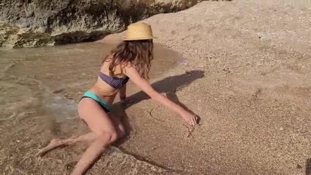 Молода худа леді в капелюсі лежить на пляжі на жовтому піску біля хвилі моря. Рука зверху намальована серцем на пляжі. Концепція літнього відпочинку подорожі — стокове відео
