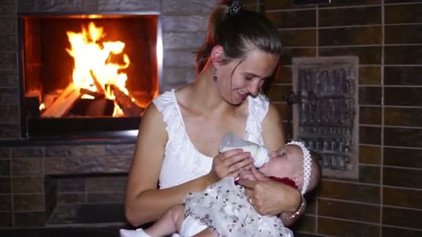 Mother feeds her baby with a bottle near a fireplace — Stock Video