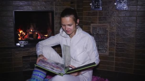 Beautiful girl sitting near the fireplace and watching photo album — Stock Video