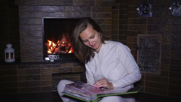 Hermosa chica sentada cerca de la chimenea y viendo álbum de fotos — Vídeos de Stock