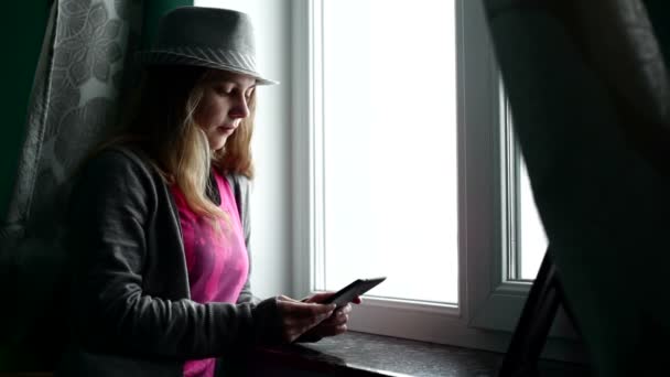 Silueta de una hermosa chica en un sombrero contra la ventana con la tableta — Vídeos de Stock