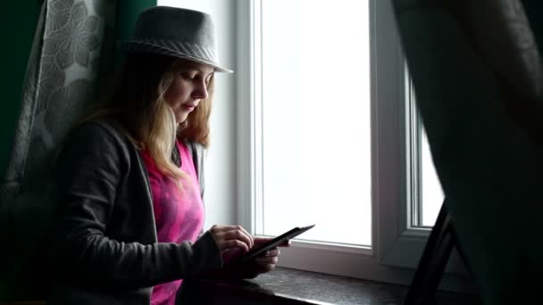 Silueta de una hermosa chica en un sombrero contra la ventana con la tableta — Vídeos de Stock