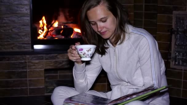 Meisje met een kopje thee bij de open haard zittend — Stockvideo