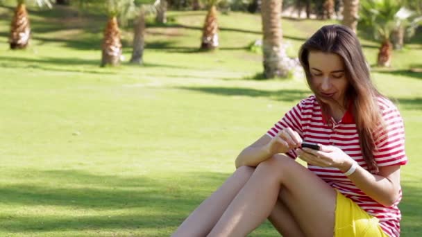 Hermosa mujer sentada en la hierba en el parque de la ciudad de verano con teléfono — Vídeo de stock