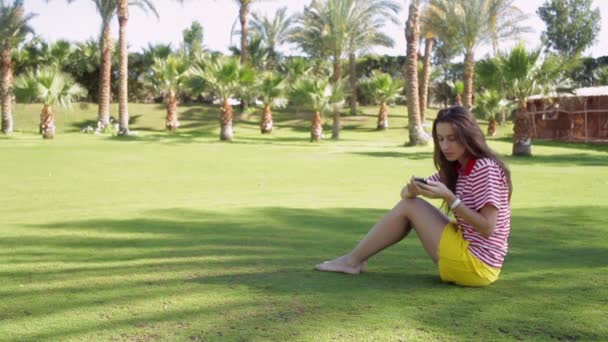 Красивая женщина сидит на траве в летнем городском парке с телефоном — стоковое видео