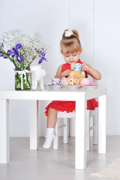 Niña en vestido rojo se sienta a la mesa y las aguas de la taza de la muñeca — Foto de Stock