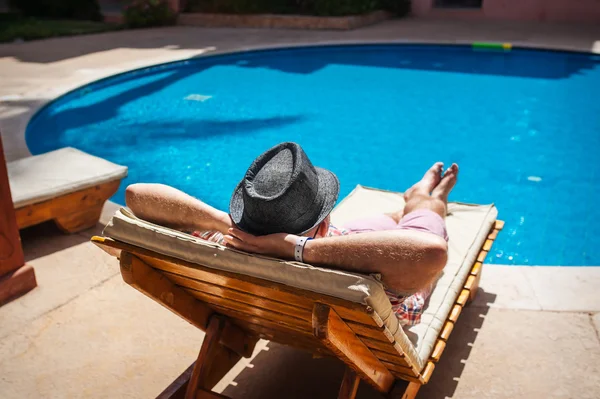 Homme au chapeau allongé sur une chaise longue près de la piscine — Photo