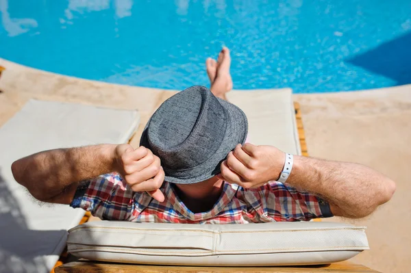 Homme au chapeau allongé sur une chaise longue près de la piscine — Photo