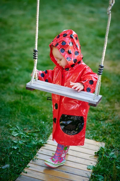 Little girl in a red jacket stands near the swing — Stok fotoğraf