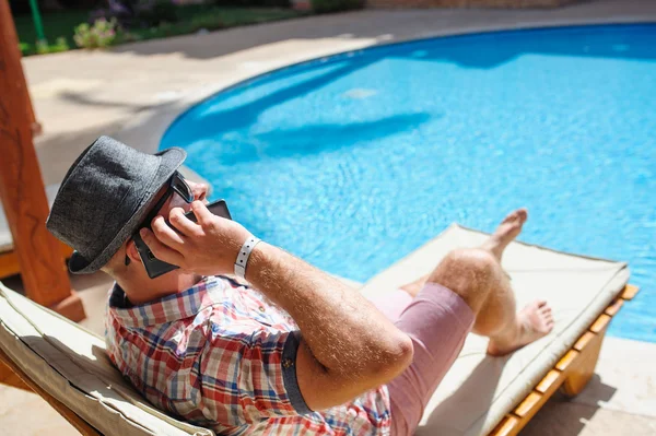 Homme au chapeau allongé sur une chaise longue près de la piscine et parlant au téléphone — Photo