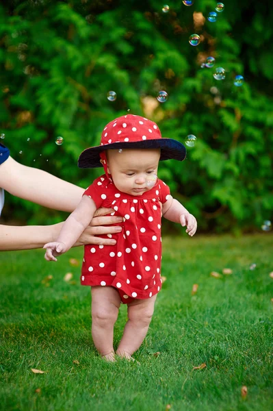 Aranyos vicces boldog baba egy piros ruhát, hogy az első lépéseket a zöld fű, egy napsütéses nyári kert — Stock Fotó
