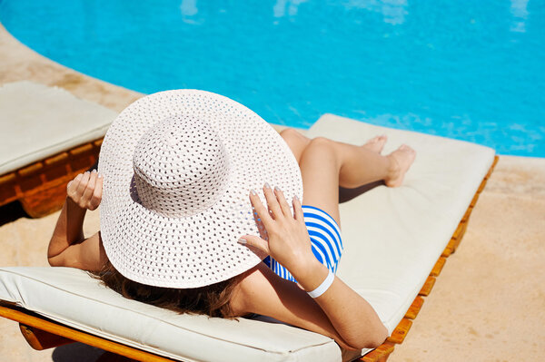 Beautiful woman in a big white hat on a lounger by the pool