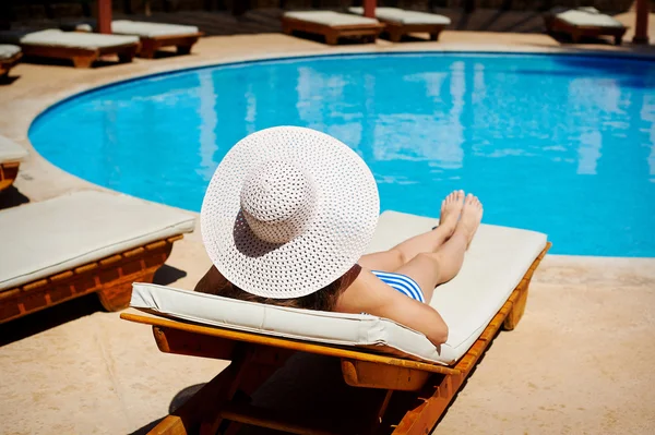 Piękna kobieta w wielkim białym kapeluszu na leżaku przy basenie — Zdjęcie stockowe
