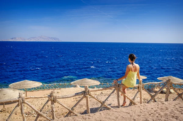 Kobieta w żółtej sukience siedząc na płocie na plaży i patrząc na morze — Zdjęcie stockowe