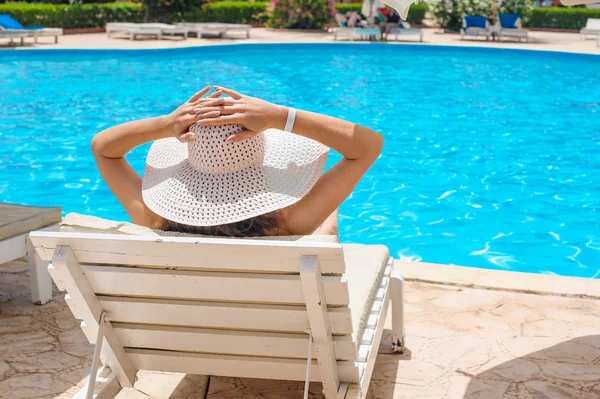 Vrouw in een grote witte hoed ontspannen op een ligstoel bij het zwembad — Stockfoto