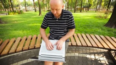 Genç adam kucağında dizüstü bilgisayarla parkta oturuyor.