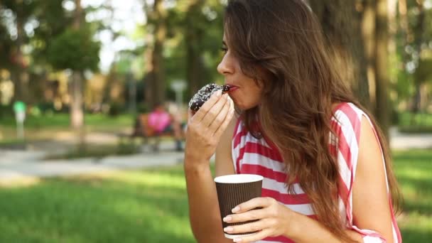 Hermosa mujer sentada en un banco en el parque y comiendo un panecillo y un café con leche — Vídeos de Stock