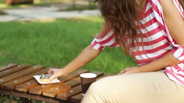 Mooie vrouw zittend op een bankje in het park en het eten van een muffin en een latte — Stockvideo