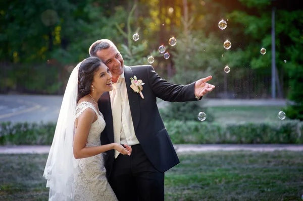 Happy bride and groom and blowing bubbles in park — Stock Photo, Image