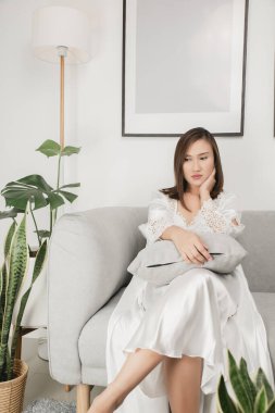 Beyaz saten uyku giysisi giyen Asyalı kadınlar gri bir kanepede yan yan bakıyor. Dalgın bir kadın.