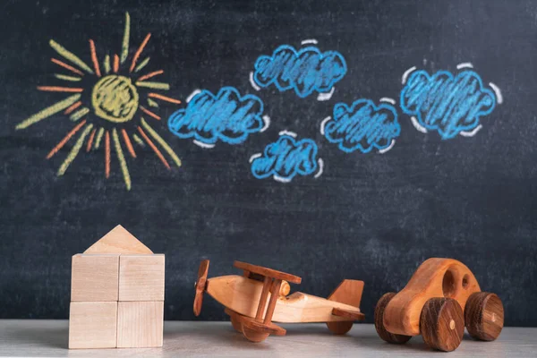 A wooden house and a toy airplane and a car stand against a chalk board with a picture of the sun and clouds. The concept of returning home from a trip