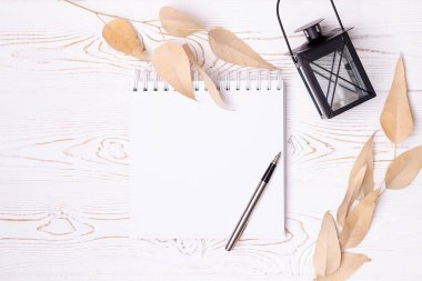 Boş bir albüm ve metal kalem, bej yapraklar, beyaz ahşap bir masada eski bir fener şeklinde siyah bir şamdan..