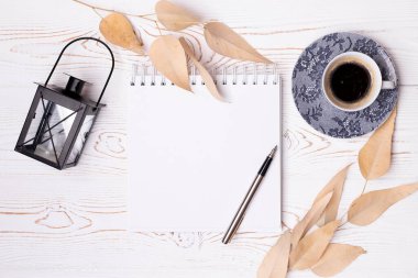 Top view of an empty scrapbook and a metal pen, a cup of coffee, beige leaves, a black iron candlestick in the form of an old lantern on a white wooden table.