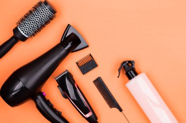 Hairdressing tools in black: hair dryer, combs, sprays, clipper on a orange background, place for text, flat lay.