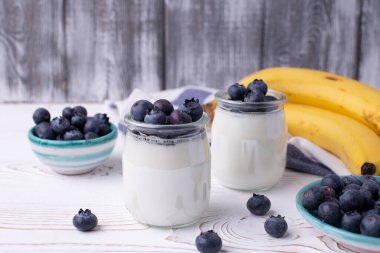 Cam kavanozlarda Yunan yoğurdu, taze yaban mersini ve kırsal beyaz bir masada olgun muzdan oluşan sağlıklı bir kahvaltı. Öndeki bulanık arka planlı cam kavanoza odaklan