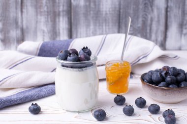 Cam kavanozda Yunan yoğurdu, yabanmersini ve kırsal beyaz bir masada reçel. Öndeki bulanık arka planlı cam kavanoza odaklan