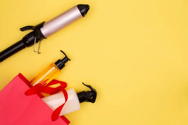 Hairdressing tools in black: curling tongs, hair brush, spray in red gift bag on yellow background, space for text, flat lay