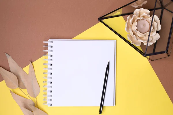 Vista dall'alto di un diario vuoto con una molla e una penna, foglie beige, florario di vetro con una candela a forma di fiore su uno sfondo combinato giallo-marrone, posto per testo Foto Stock