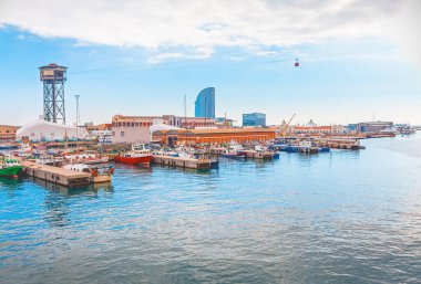 Barcelona 'daki Port Vell manzarasında rıhtımlı küçük tekneler, tarihi rıhtım binaları ve arka planda kendine özgü Torre Jaume I yer almaktadır. Şehrin ufuk çizgisi bulutlu bir gökyüzünün altında görünür.