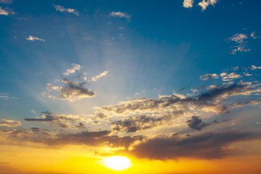 Golden sunlight pierces scattered clouds as sky shifts from fiery orange to tranquil blue, capturing the serene drama of a transitional moment. Rays fan outward, adding depth and quiet intensity