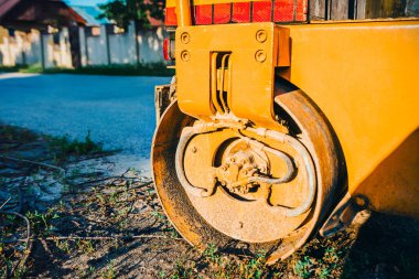 Yellow road roller with a large, rusty metal drum resting on a patch of grass and dirt. Freshly paved asphalt road is visible in the background, hinting at recent construction