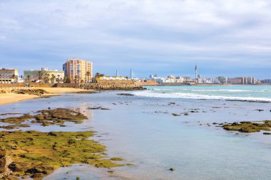 İspanya 'nın Cadiz kentindeki kıyı şeridi manzaralı, özellikle Playa de la Caleta yakınlarında. Sahilin eşsiz kayalık ön planı, turkuaz su ve parlak bir gökyüzünün altındaki uzak şehir manzarası.