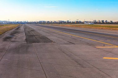 Beton ve asfalttan yapılmış, sarı işaretli bir havaalanı pisti manzarası. Sahne, uzaklarda birkaç uçak, uzak bir şehir silueti ve açık mavi gökyüzü içerir.