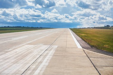 Geniş beton pist, parçalı bulutlu bir gökyüzünün altında çimenli tarlalarla çevrili olan mesafeye kadar uzanıyor. Beyaz işaretler uçak kalkış ve iniş için yol gösteriyor, ufukta görünen uzak binalar var.