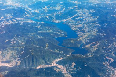 Hava görüntüsü ormanlık tepeleri ve vadileri kesen dolambaçlı mavi bir su deposu gösteriyor. Yamalı arazi ve uzak yerleşim alanları açık gün ışığında geniş ve katmanlı bir manzara yaratır.