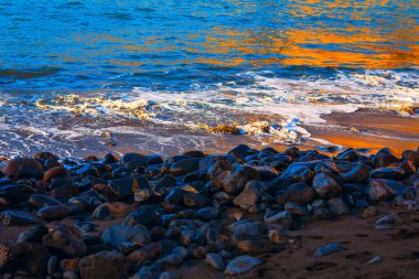 Altın ışığın, koyu ve pürüzsüz taşlarla kaplı kıyılarla buluşurken nazik okyanus dalgalarını yansıttığı huzurlu bir an. Parlak mavi su ve gölgeli kayalar arasındaki zıtlık dramatik bir doğal manzara yaratır.