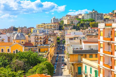 Densely packed cityscape on steep hill, likely in Italian city of Messina, Sicily. Scene is filled with traditional Mediterranean buildings, narrow streets, and greenery under blue sky