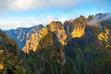 Portekiz, Madeira 'daki çarpıcı Pico do Arieiro dağ manzarası, sivri tepeler ve bulutların üstündeki yürüyüş patikaları. Sabah güneşi sert kaya yüzeyleri ve adanın yeşil yamaçlarını çarpıcı bir şekilde vurguluyor.