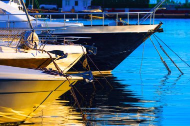 Yat yayı bir marinaya kenetlenmiş, gövdeleri sıcak günbatımı ışığı ile parlıyor ve sakin mavi suda yansıtılıyor. Zincirler ve ipler onları güvenli bir şekilde bağlarken, binalar da sakin deniz manzarasını çerçeveliyor.
